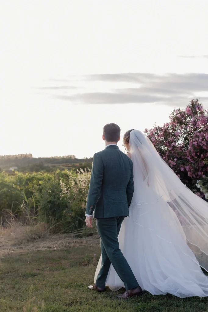 Mathilde et Romain marchent de dos au Mas des Tourelles, le long voile de la mariée flottant derrière eux